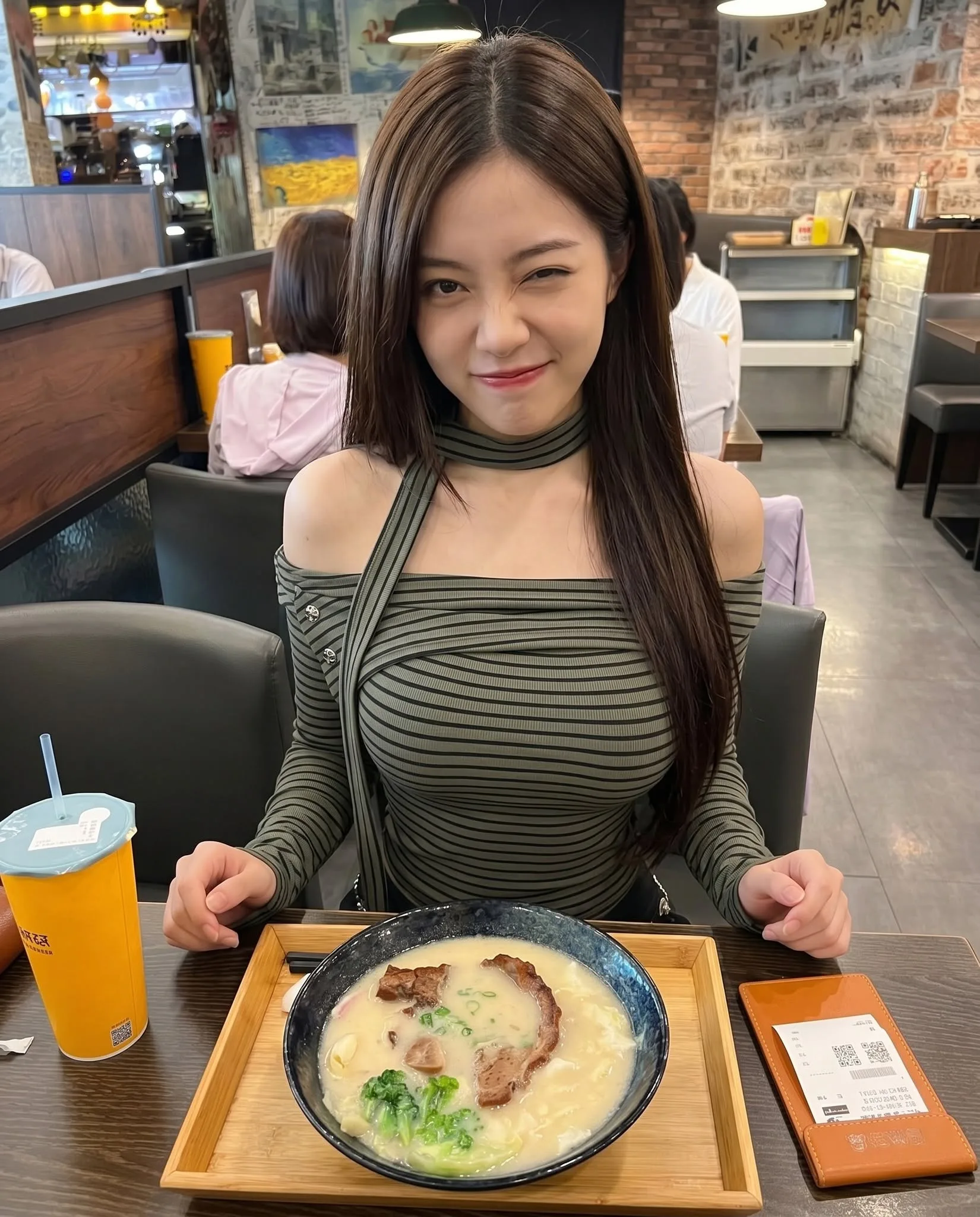 一位留著長直髮的年輕女性，身穿綠色條紋露肩上衣，在餐廳對著鏡頭俏皮眨眼，桌上擺著一碗拉麵與飲料。